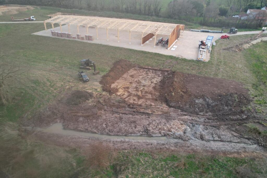Vue aérienne d'un chantier de terrassement avec une tranchée creusée pour la pose de câbles ou de canalisations, sur un chemin en pleine nature, avec une mini-pelle au fond du tracé.